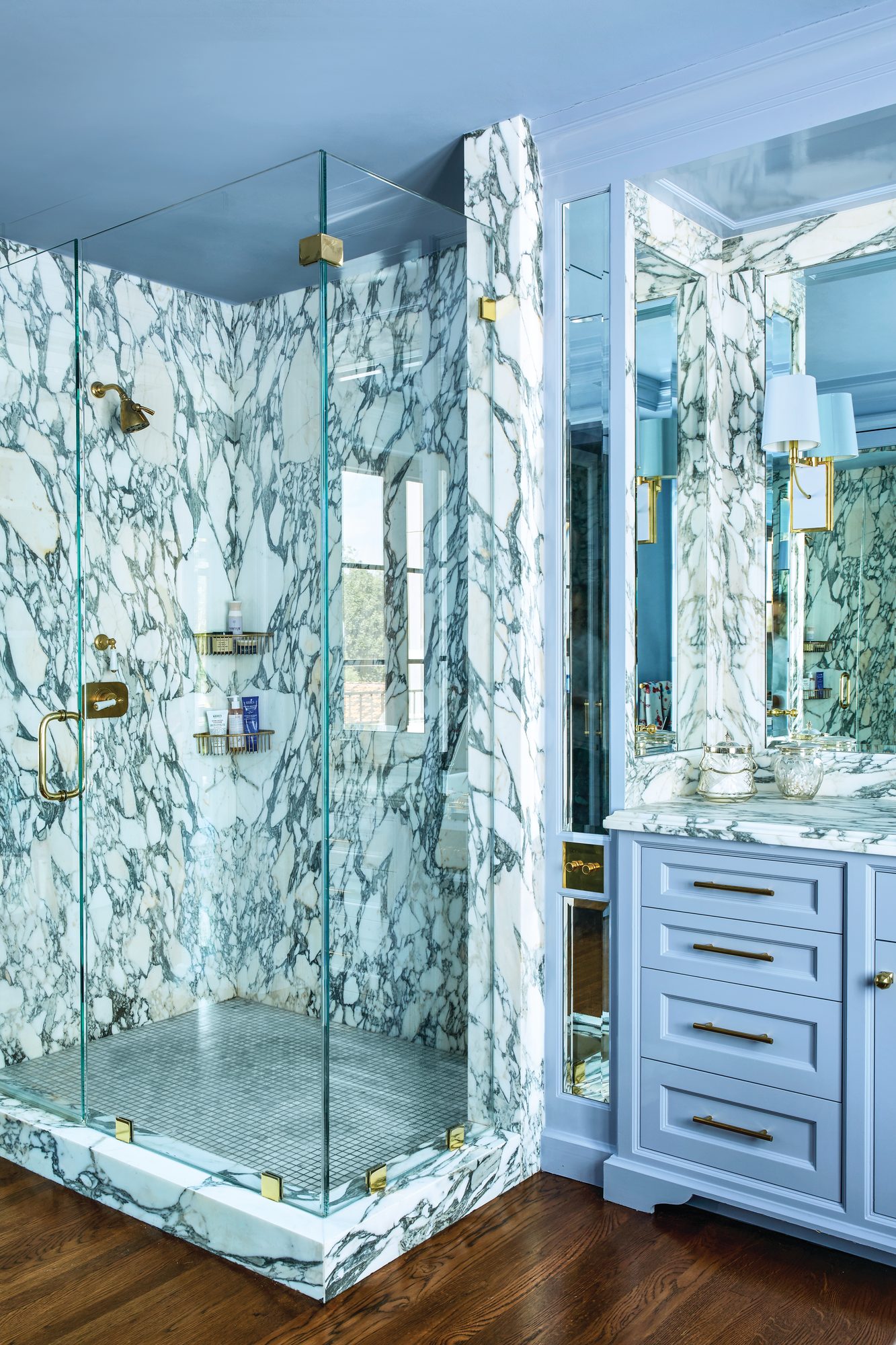 Primary bathroom with sky blue walls and cabinetry, a glass shower clad in bold veined marble, brass fittings, and mirrored panels
