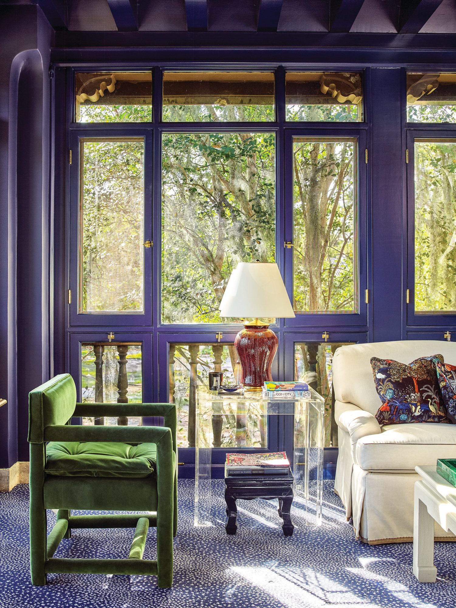 Sitting area with deep blue walls and windows, a green velvet square armchair, acrylic side table, red lamp, and blue animal print carpet