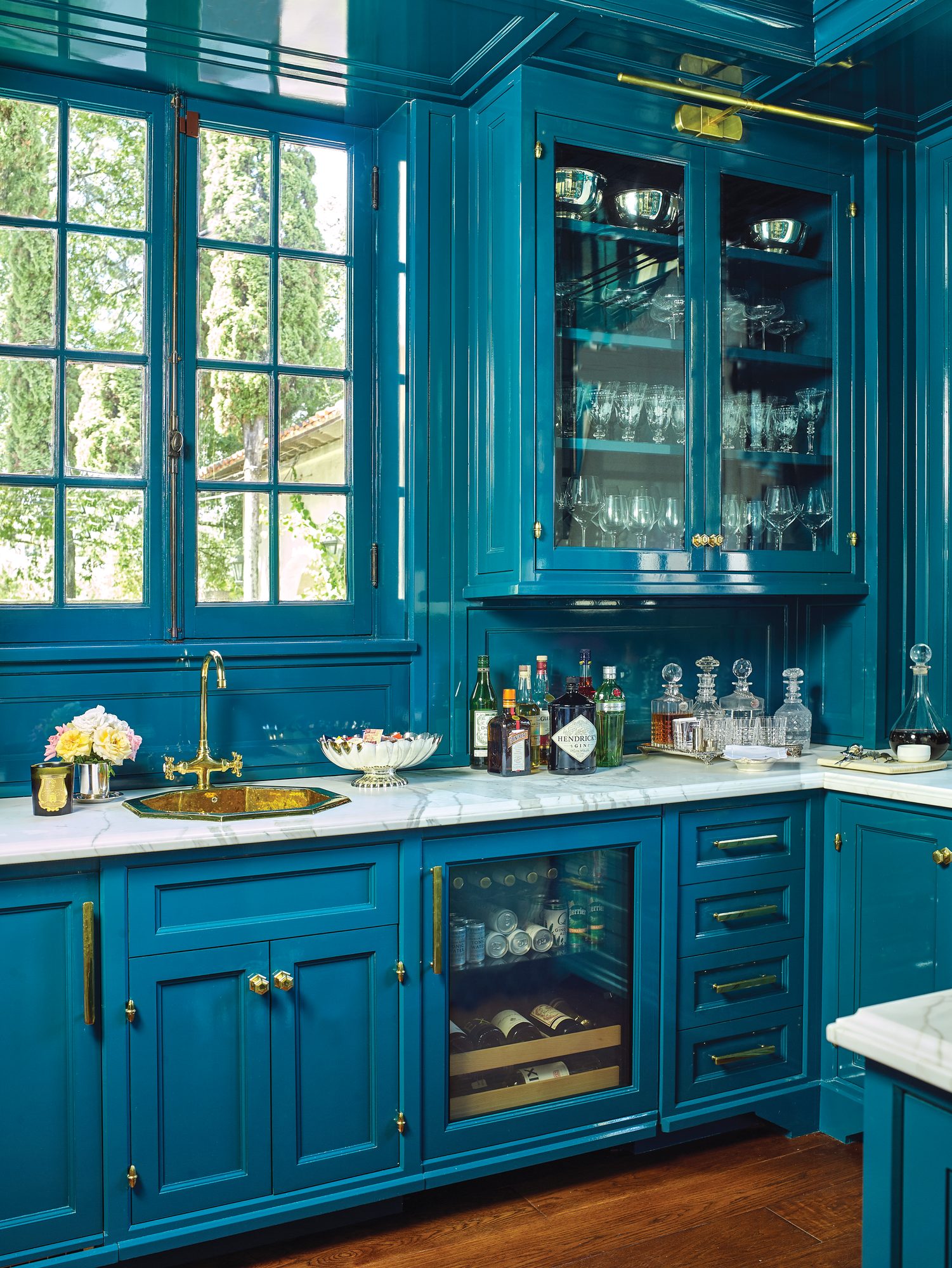 Bar with lacquered dark turquoise cabinetry, glass-front upper cabinets, brass hardware, a brass sink, marble counters, and a wine refrigerator