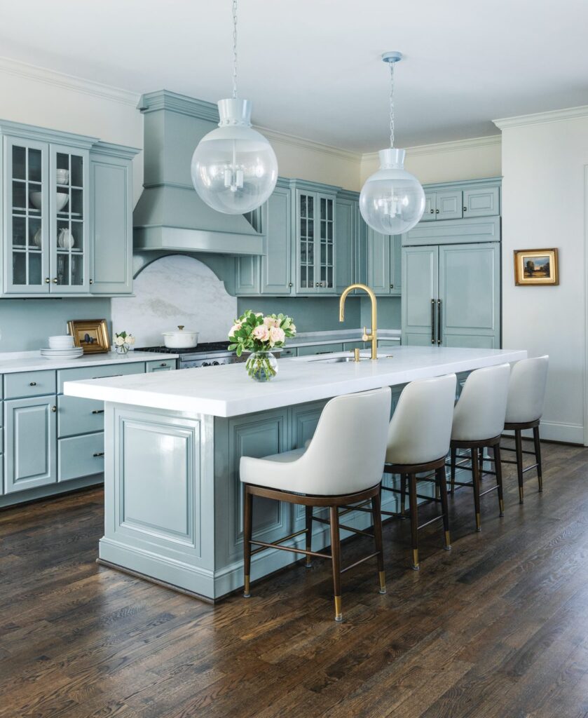 Kitchen with pale aqua cabinetry and range hood, a marble-topped island with white leather counter stools, and glass globe pendants