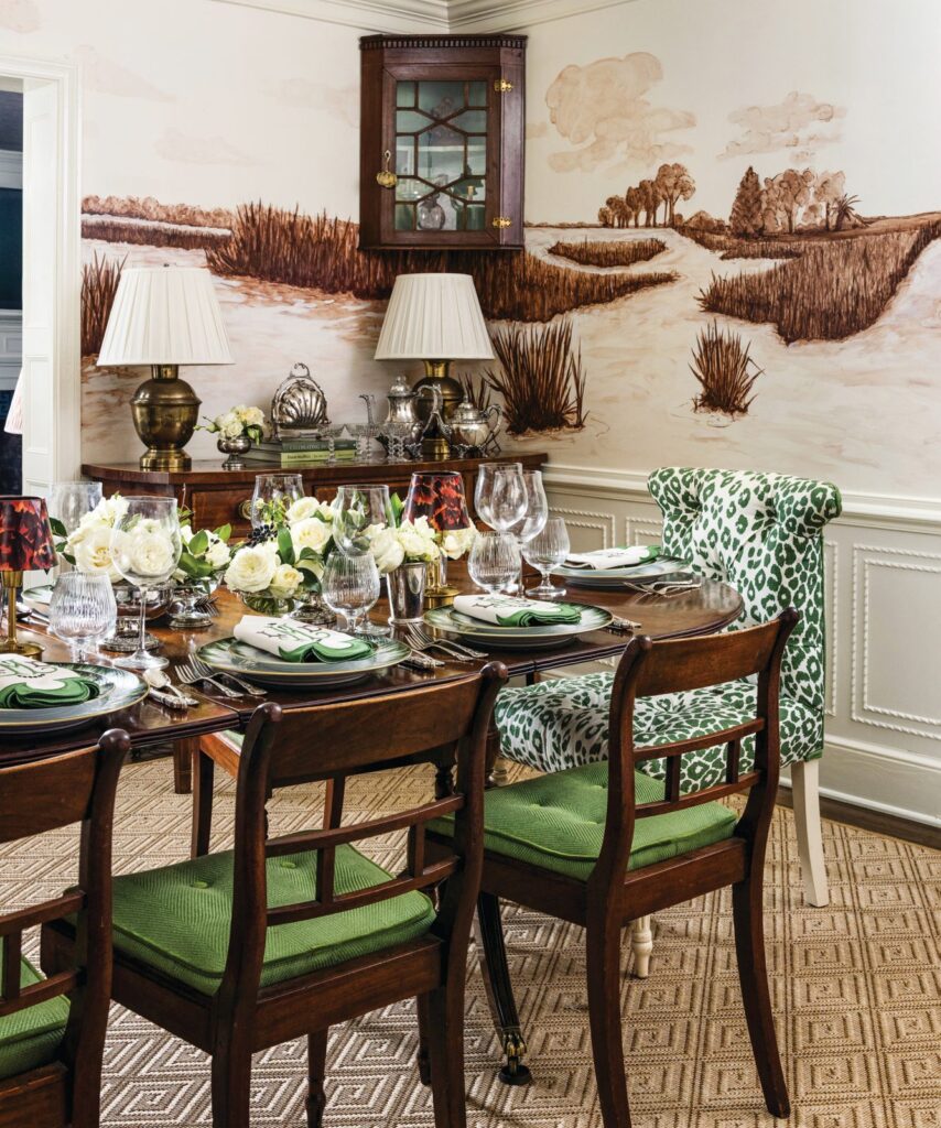 Dining room set for a party with a sepia landscape mural, green-upholstered chairs, white flowers, and crystal glassware