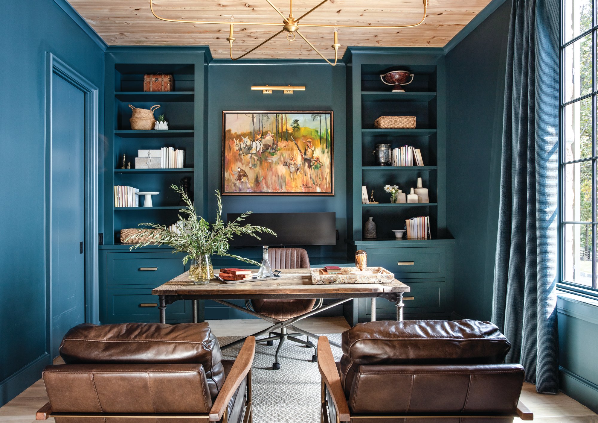 Study color-drenched in deep teal blue with built-in bookcases, a reclaimed wood desk, brown leather chairs, and a commissioned hunting painting