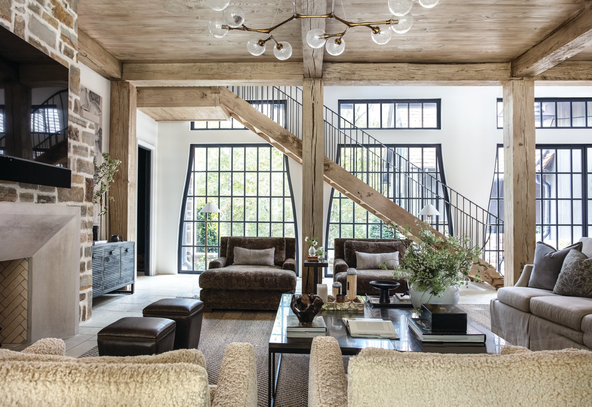 Great room with reclaimed timber columns and beams, a staircase with floating treads, black steel windows, and a stone fireplace with a cast stone mantel