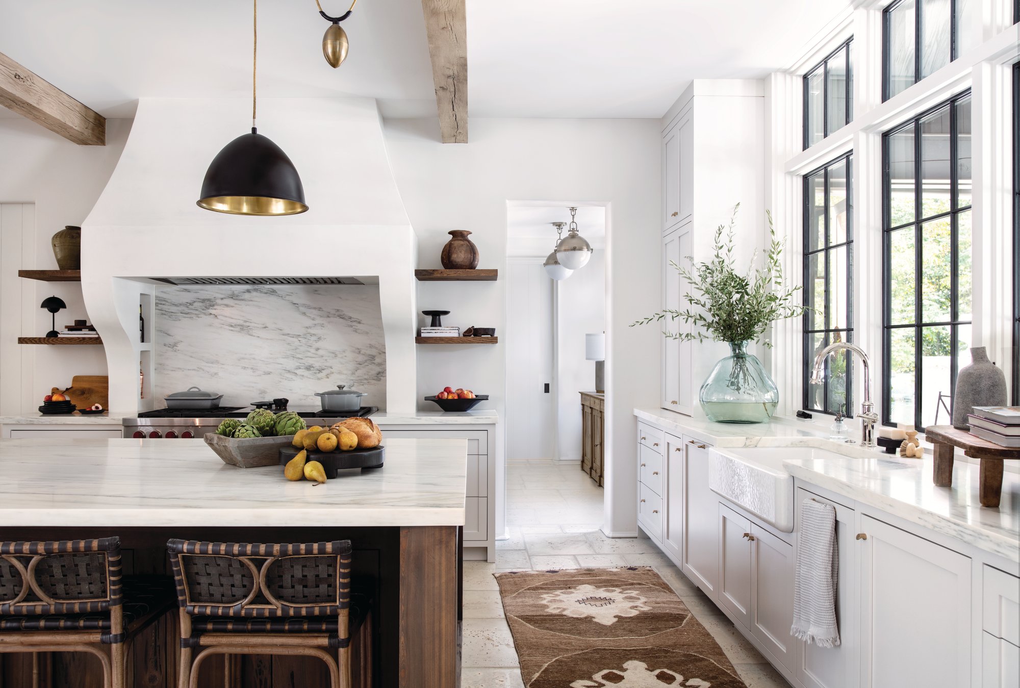 White kitchen with a plaster range hood, marble backsplash and island, a farmhouse sink under black steel windows, and woven leather counter stools