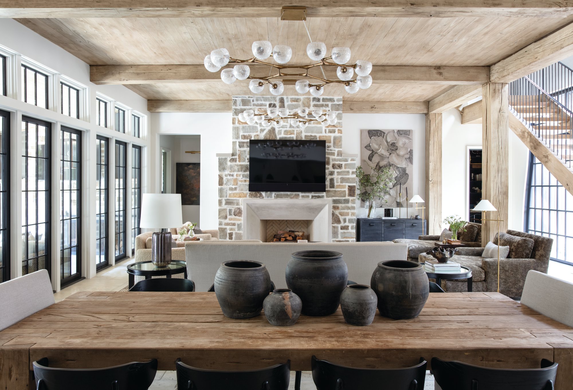 Open dining area and great room with a reclaimed wood table, stone fireplace, timber beams, and a commissioned magnolia painting by Emily Morgan Brown