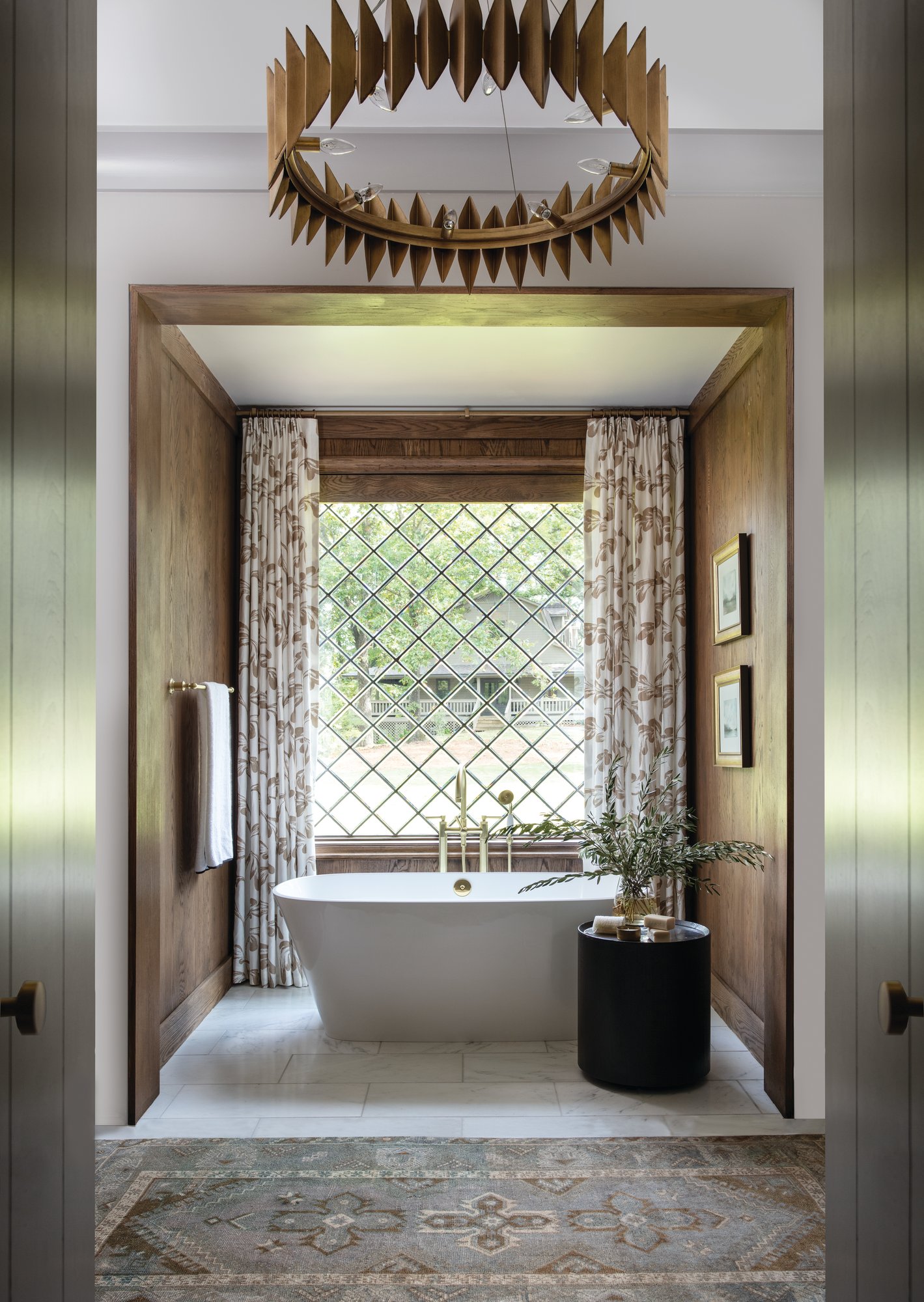 Primary bath tub nook lined in white oak paneling with a freestanding tub, a leaded glass window, and a contemporary brass chandelier