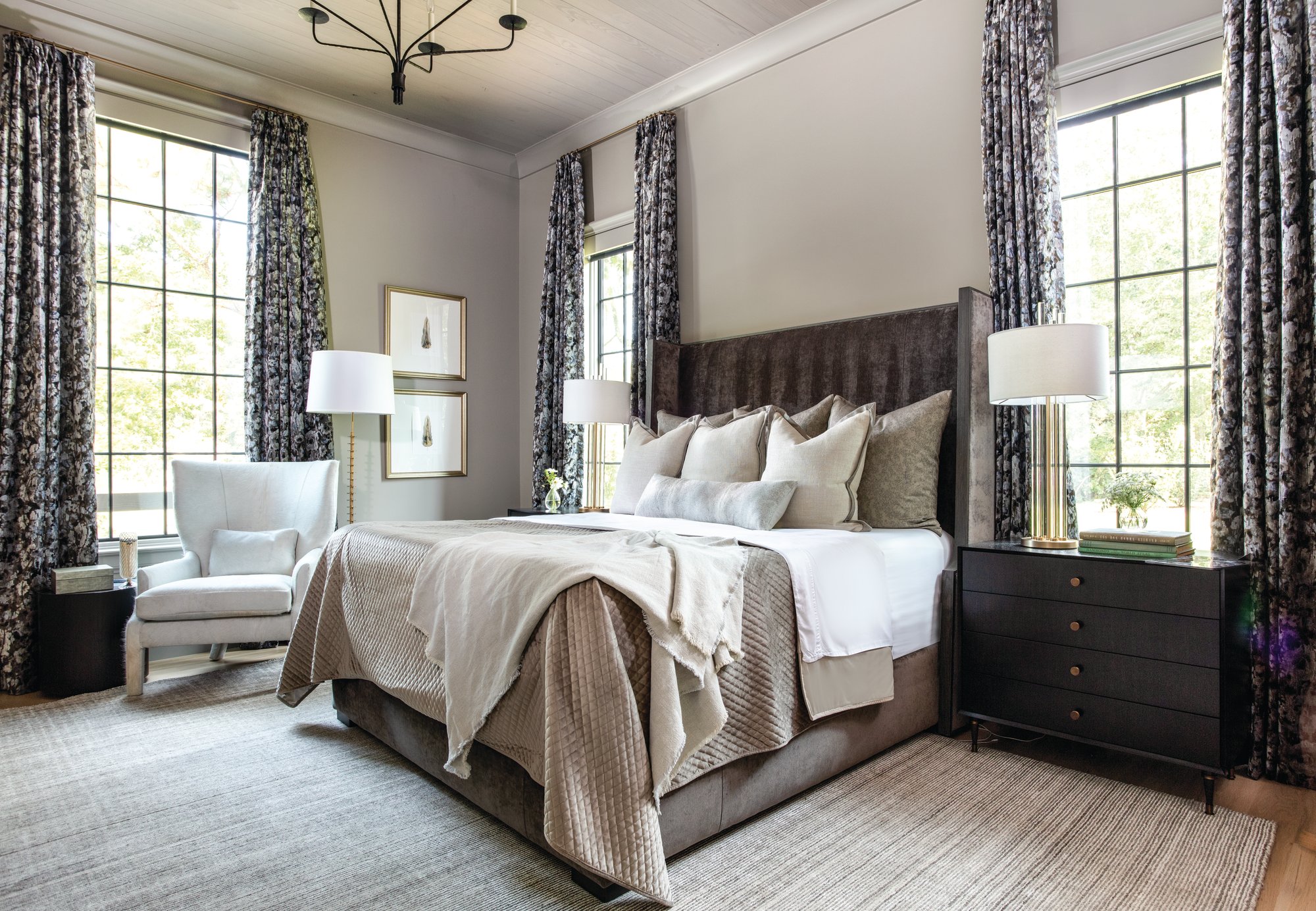 Primary bedroom with a charcoal velvet upholstered bed, floral drapery, dark wood nightstands, and a white wingback chair