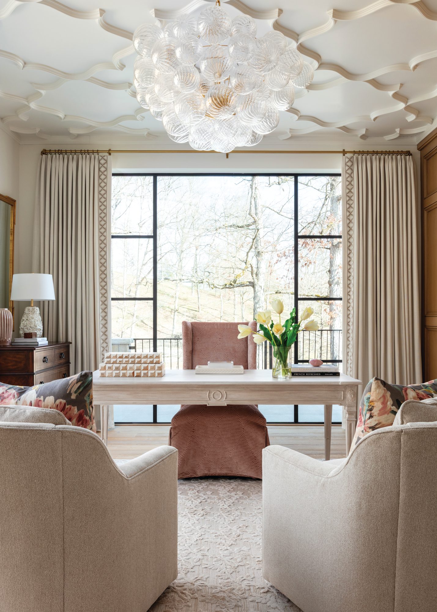 Study with a sculpted applied-moulding ceiling, glass bubble chandelier, pale desk facing a black-framed window, and a pink desk chair
