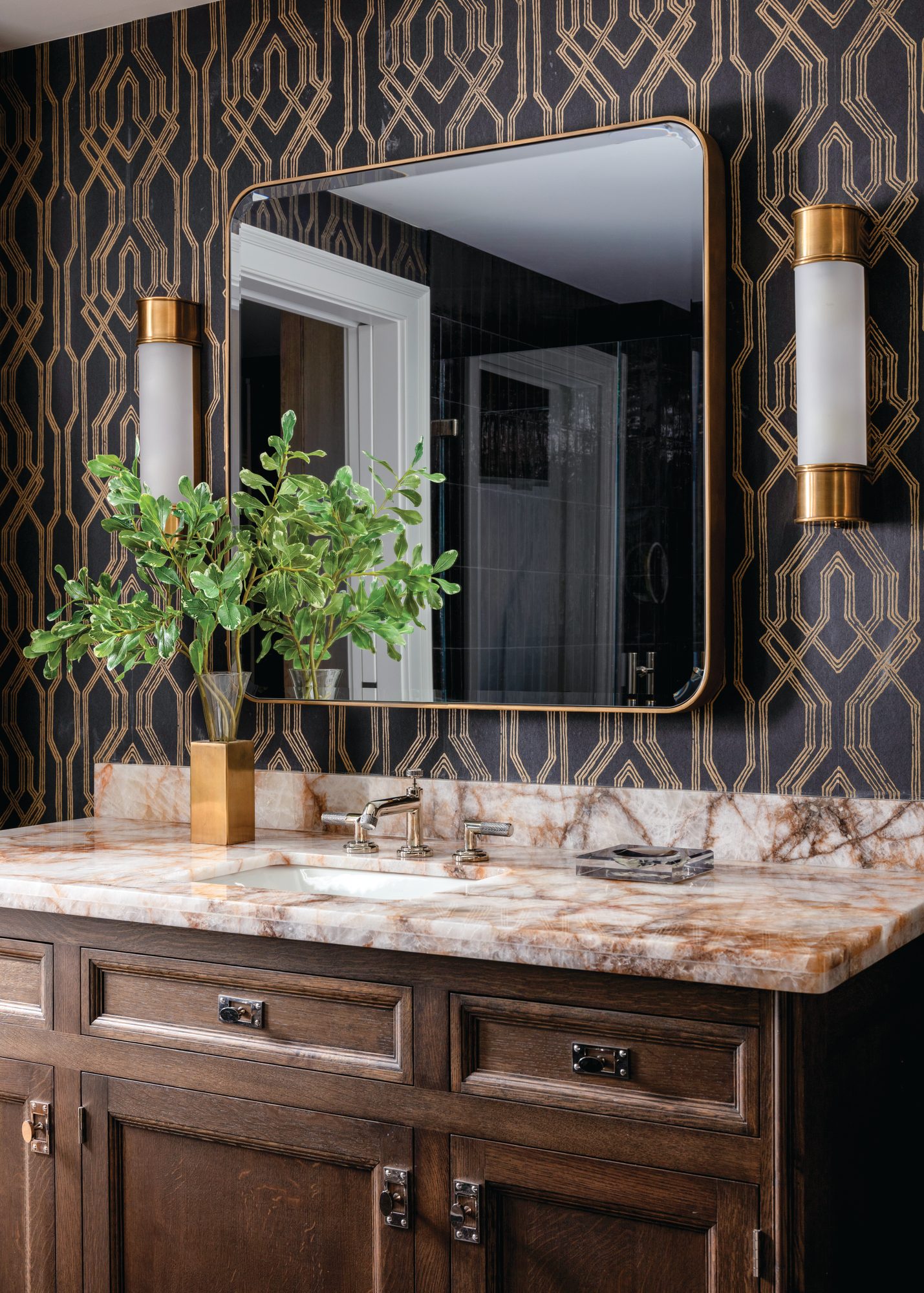 Dark-stained wood vanity with a veined stone countertop, brass sconces, and navy-and-gold trellis wallpaper in a masculine bath