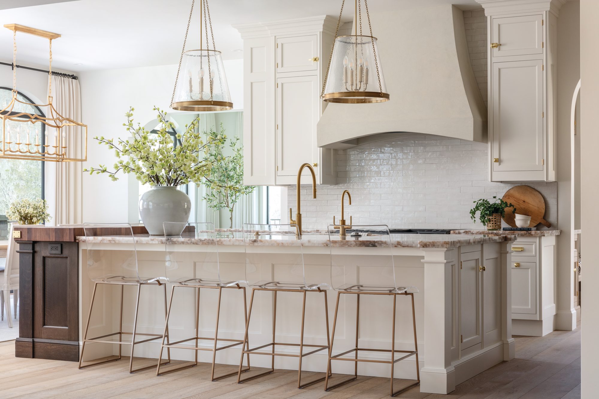 Neutral kitchen with a large stone-topped island, brass lantern pendants, a plaster range hood, and mixed painted and stained cabinetry