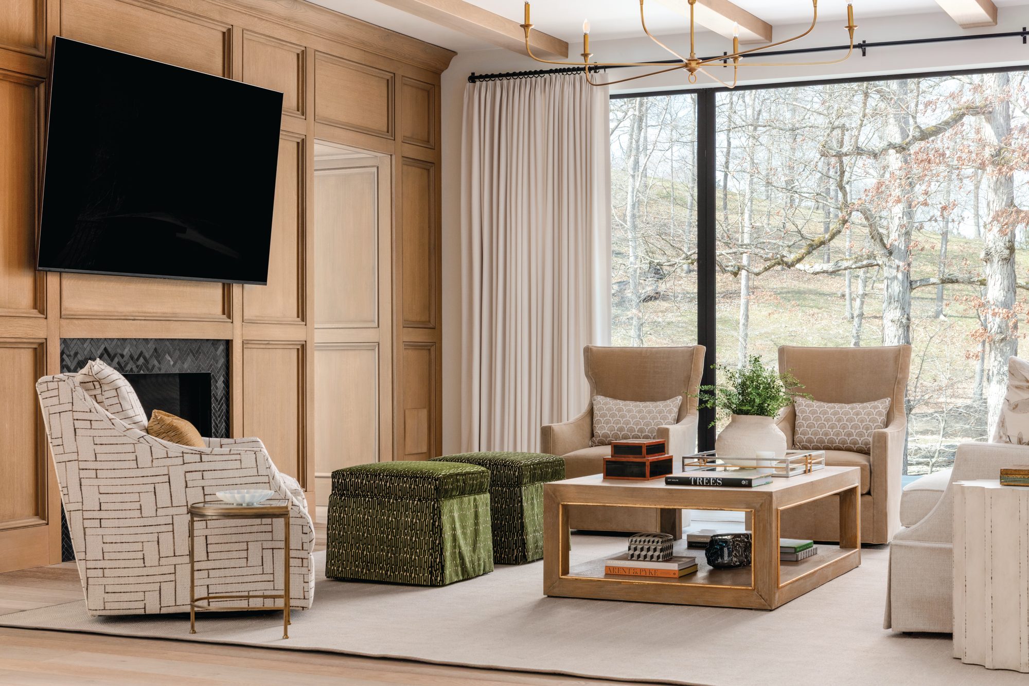 Living room with wood-paneled walls, a fireplace, green upholstered ottomans, and floor-to-ceiling windows overlooking the wooded property