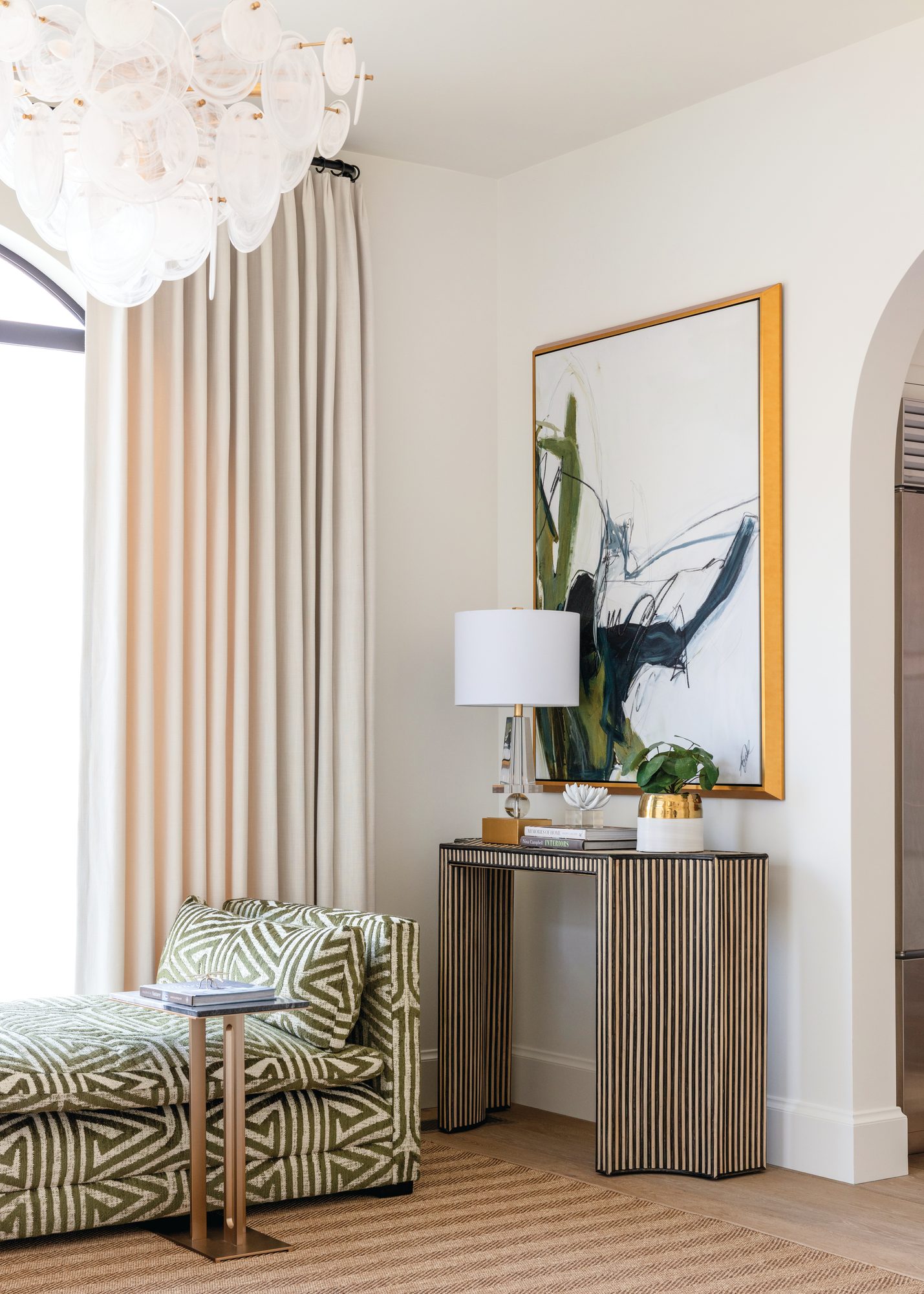 Green geometric-patterned daybed beside a fluted console table under abstract green-and-black artwork and a glass bubble chandelier