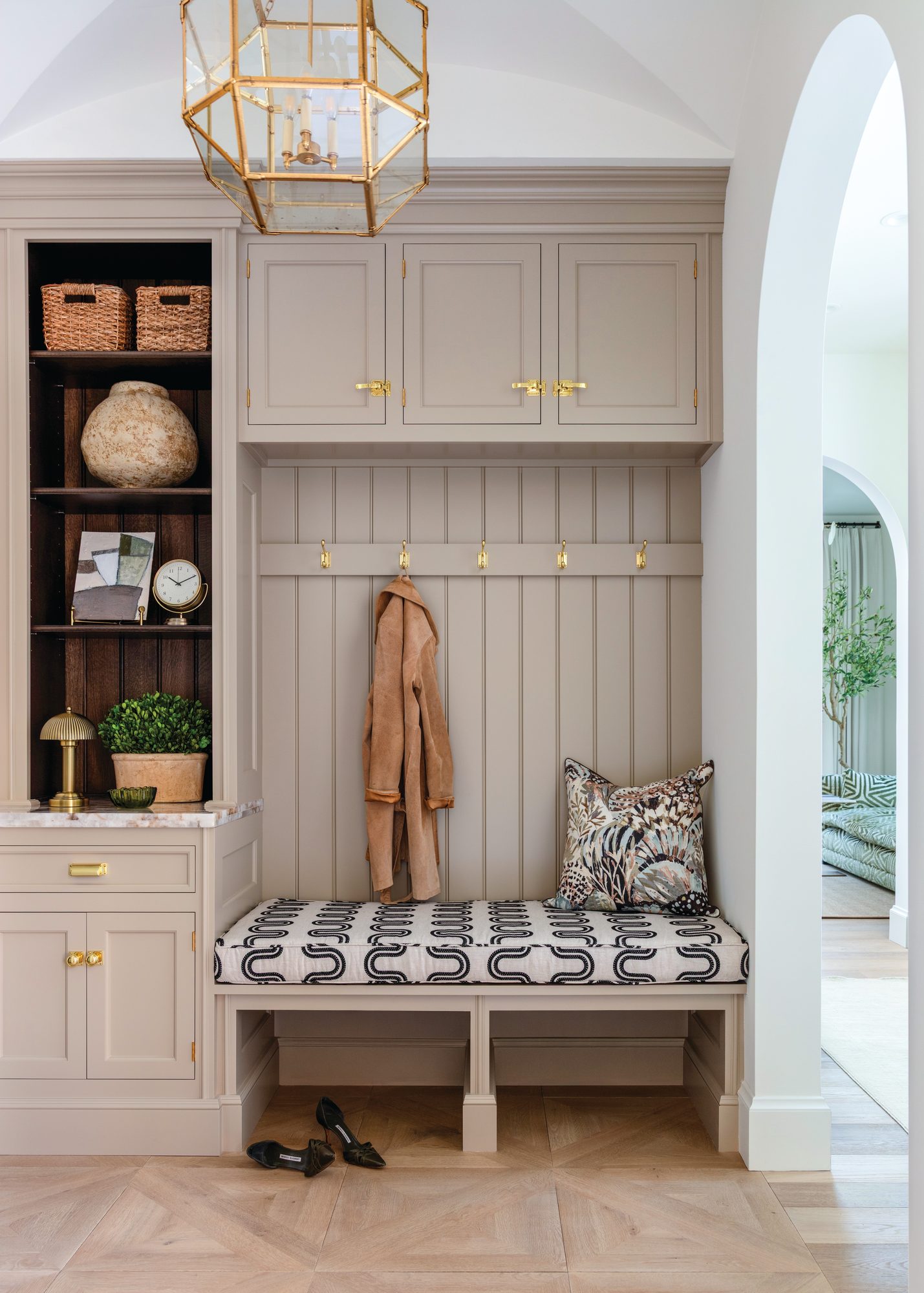 Drop zone with beadboard paneling, brass coat hooks, a cushioned built-in bench, and upper cabinetry above a herringbone wood floor