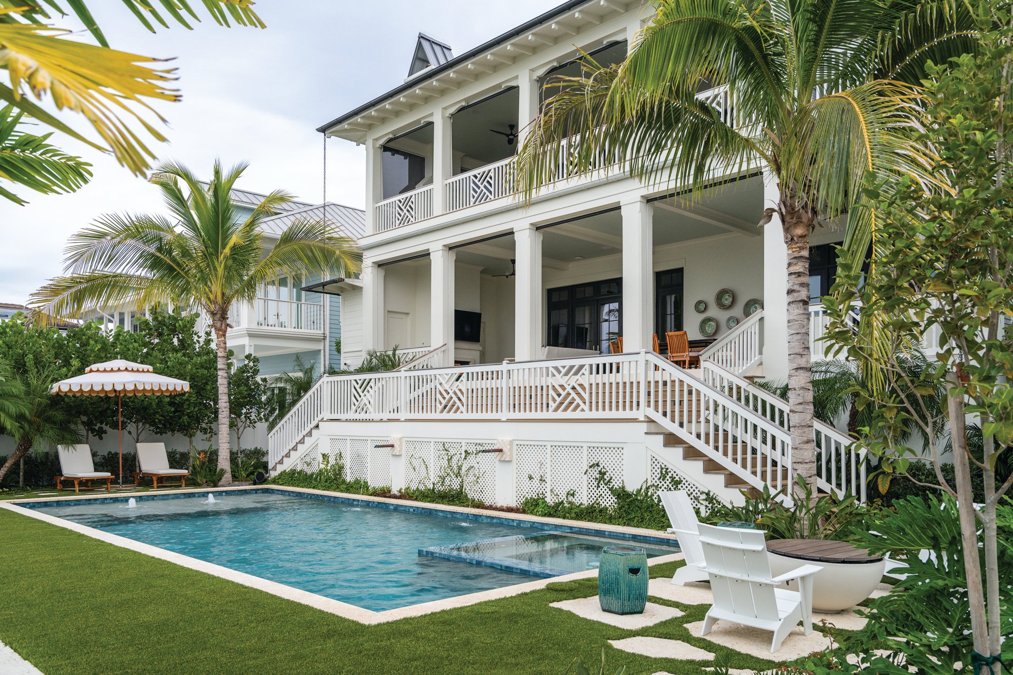 Back exterior of a white waterfront home with stacked two-story porches, Chippendale railings, palm trees, and a rectangular pool with lounge chairs