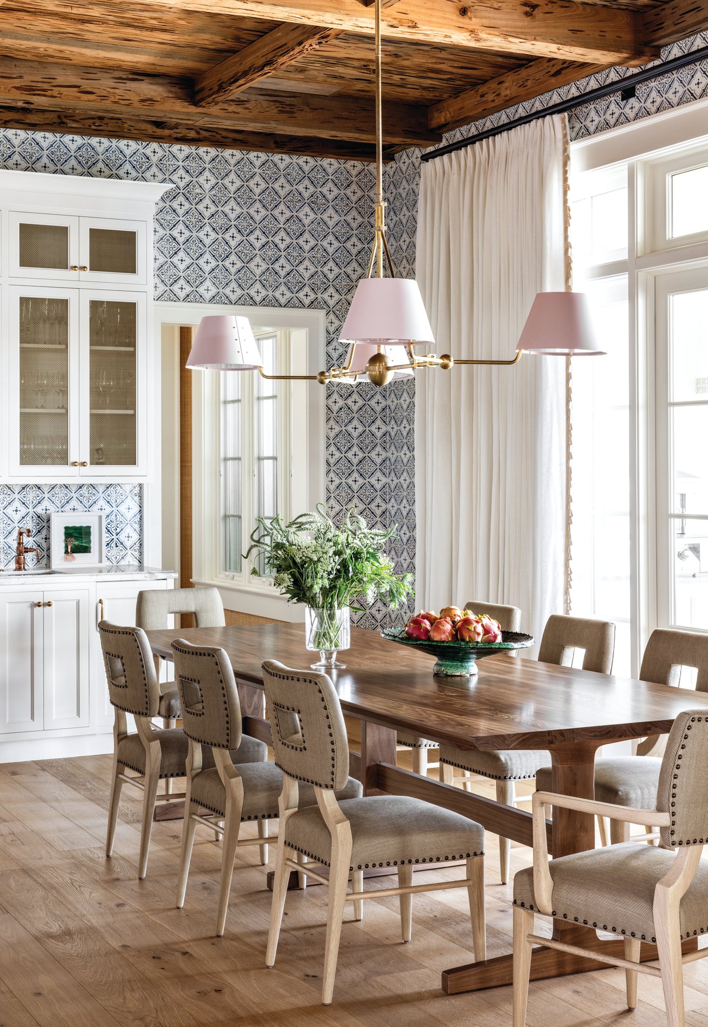 Dining area with a walnut table, nailhead-trimmed upholstered chairs, a blue and white patterned tile wall, pecky cypress beams, and a pink-shaded brass chandelier
