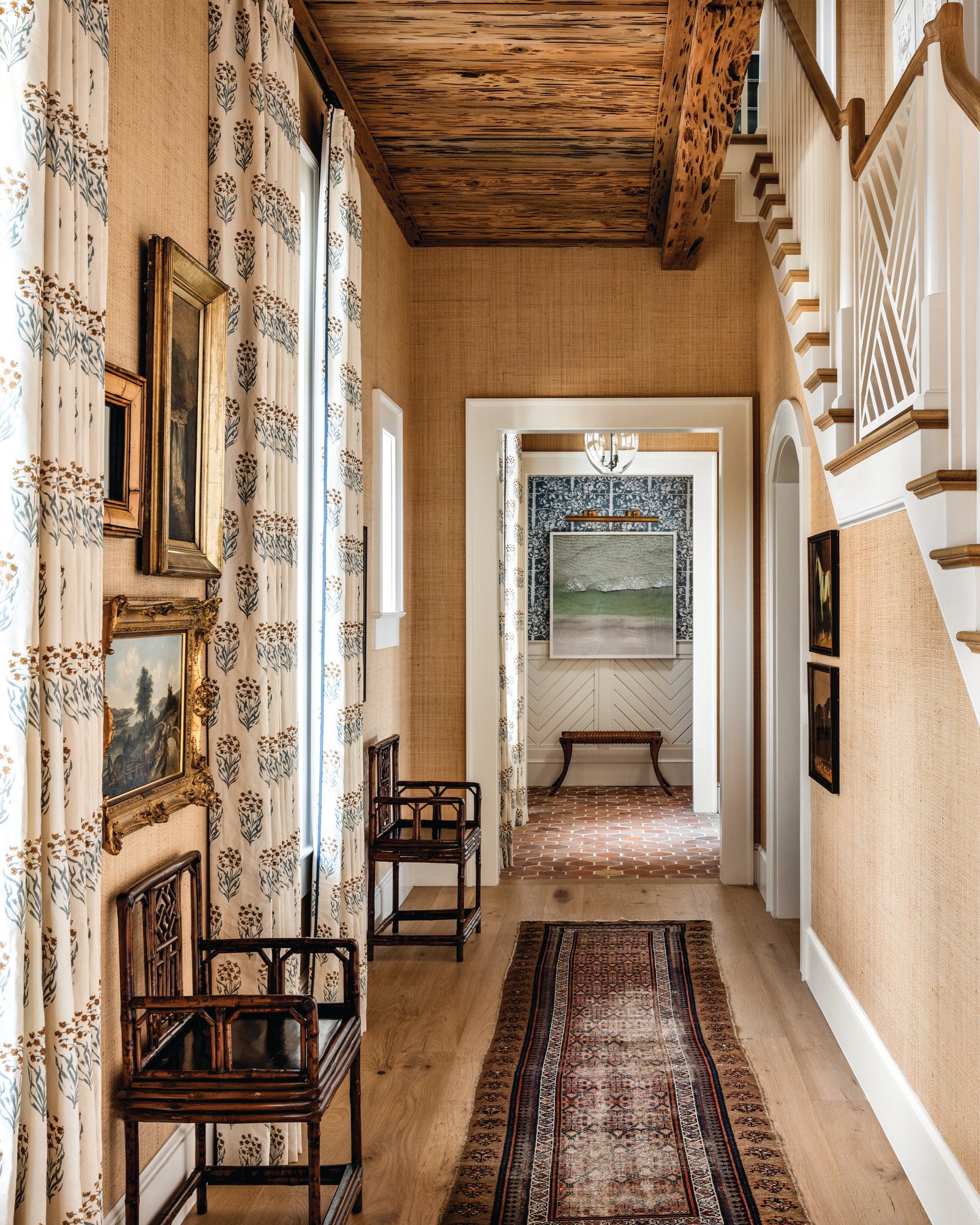 Entry hall lined in grasscloth with gilt-framed antique oil paintings, bamboo chairs, a pecky cypress ceiling, an antique runner, and a white oak staircase