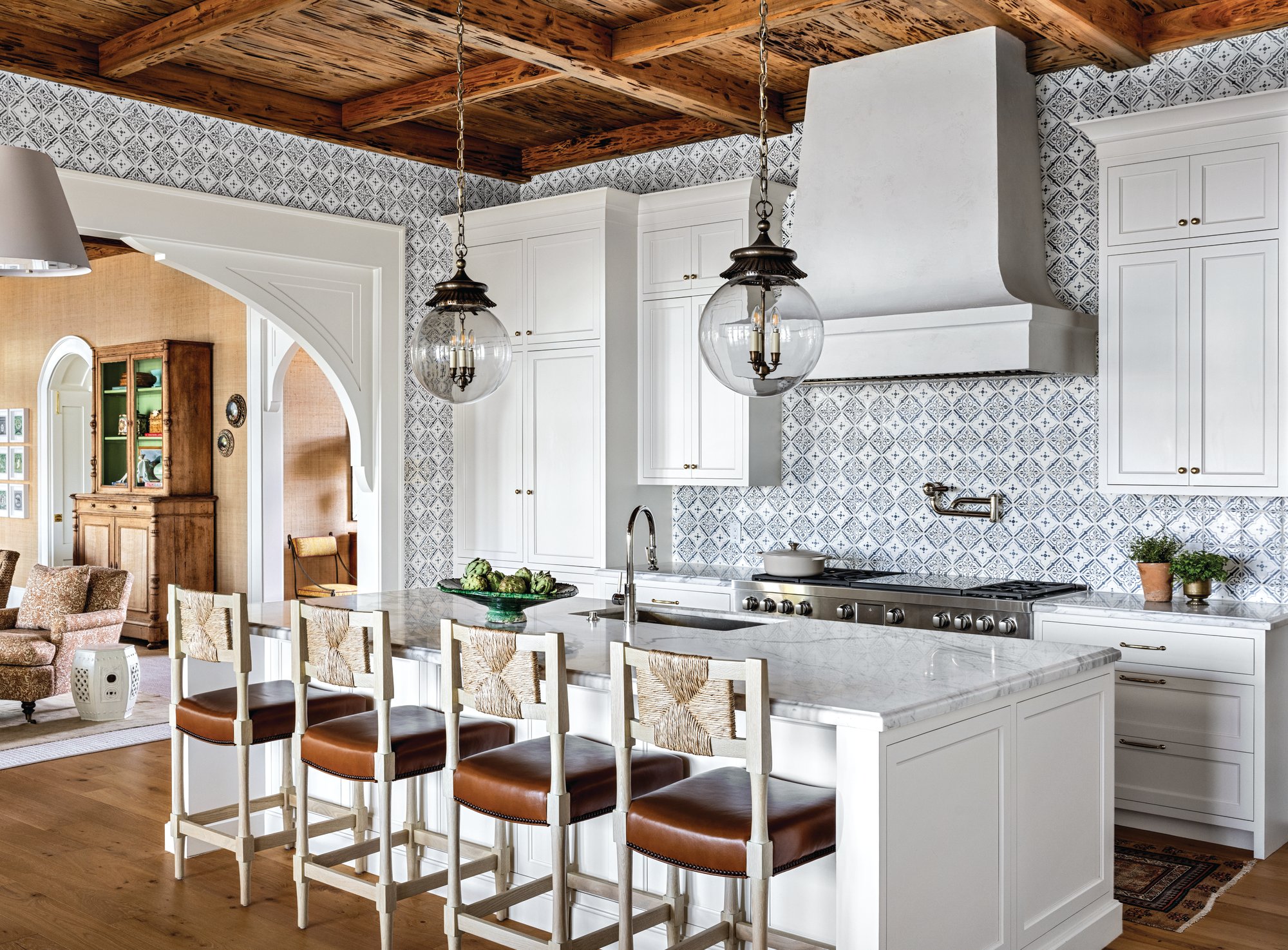 Kitchen with blue and white patterned tile walls, a plaster range hood, white cabinetry, a marble island, and woven-back leather counter stools