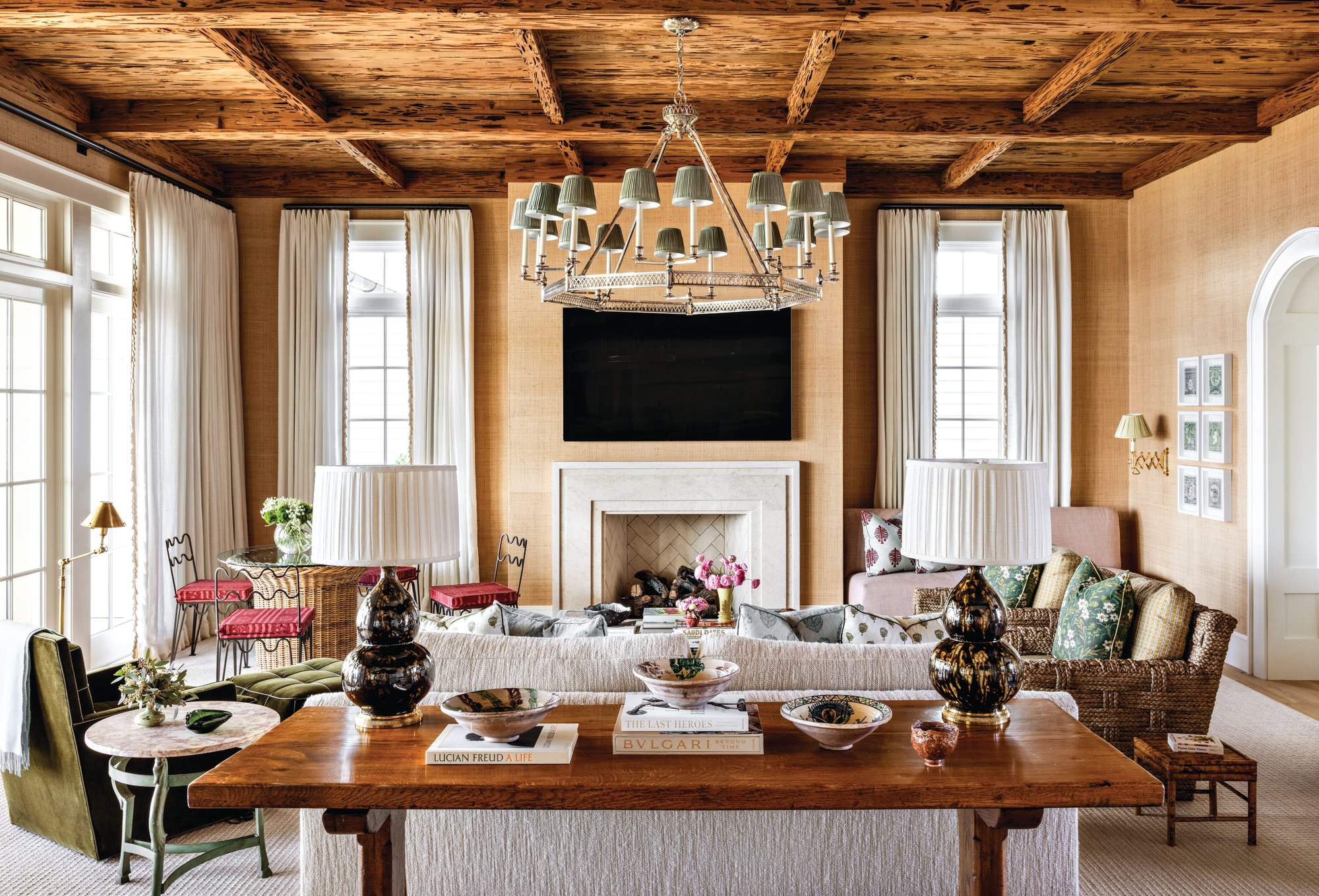 Living room with a coffered pecky cypress beam ceiling, grasscloth walls, a limestone fireplace, a silver chandelier, and layered antique and woven furnishings
