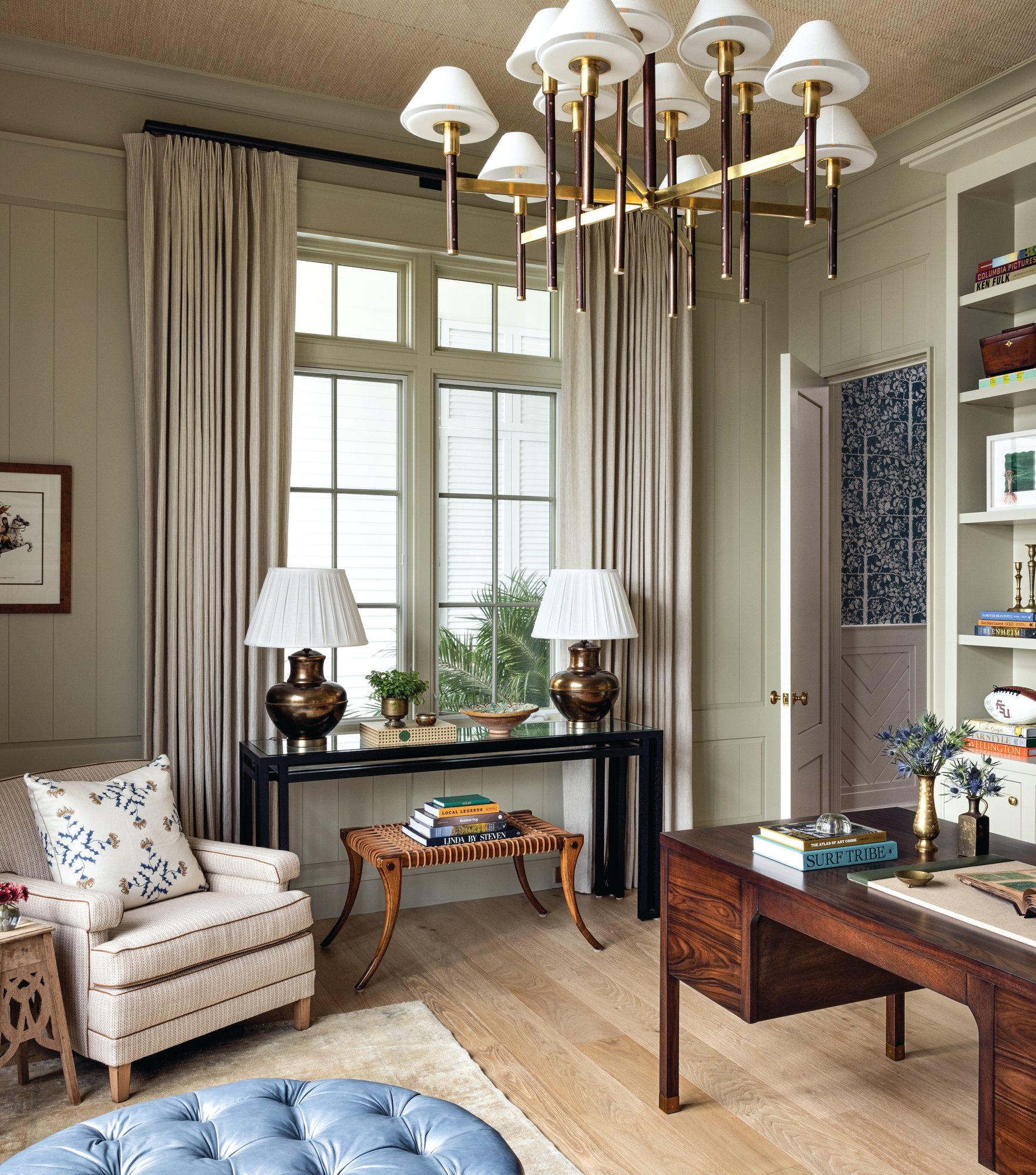 Home office with sage paneled walls, a grasscloth ceiling, a brass and leather chandelier, an antique wood desk, built-in bookshelves, and a blue tufted ottoman