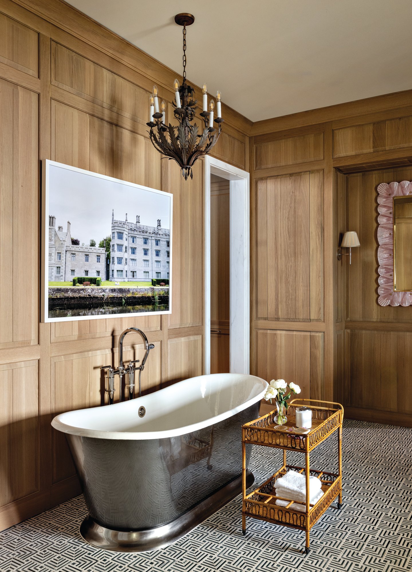 Primary bath with white oak paneled walls, a polished metal soaking tub, graphic mosaic floor tile, an iron chandelier, and a large framed travel photograph